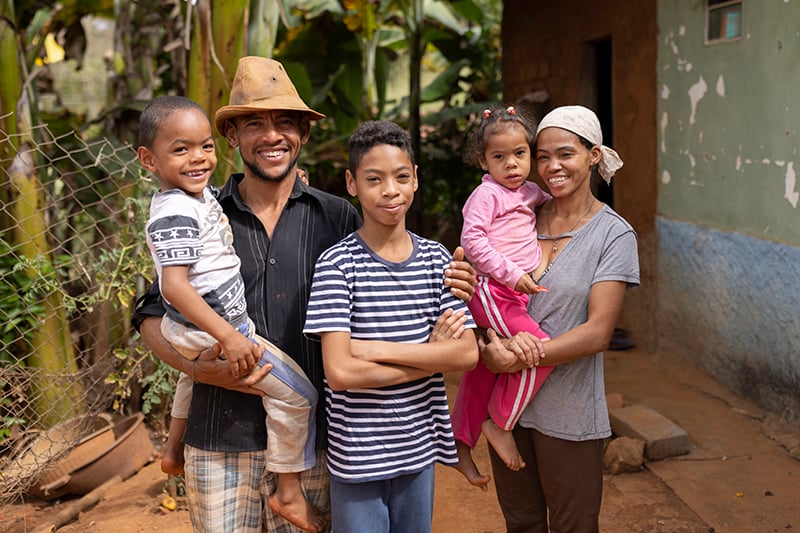 A smiling family of four poses outside their rural home, with the father holding a young boy and the mother holding a little girl.