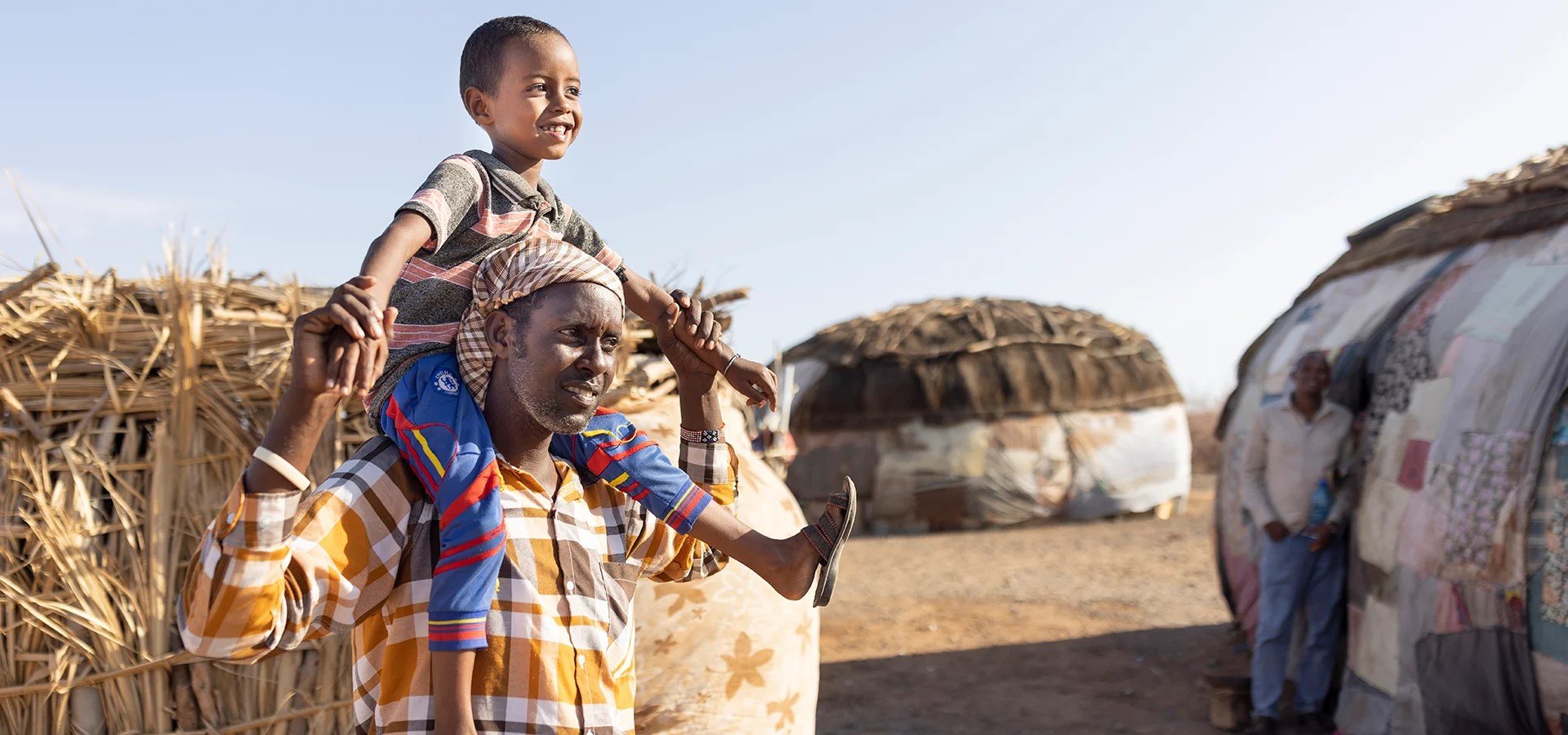 A smiling young boy sits on a man's shoulders, holding his hand, in a rural village setting with traditional round huts in the background.