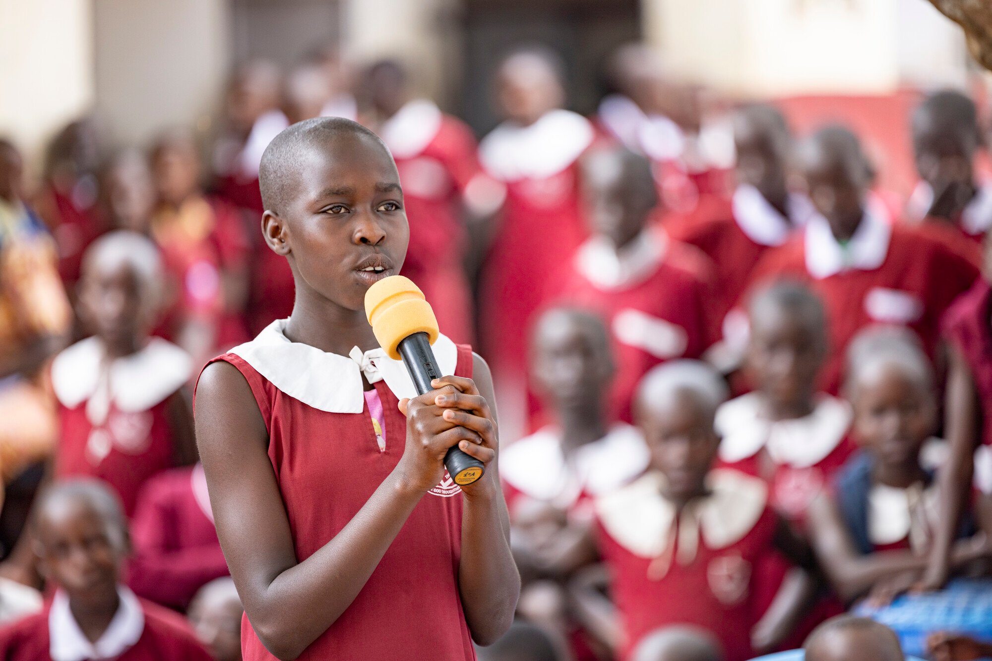 A young girl in a red and white school uniform holds a microphone and speaks to a group of students in matching uniforms in the background.