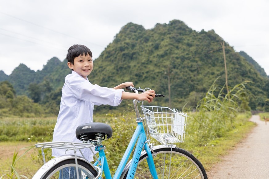 A smiling boy in a white shirt stands next to a blue bicycle with a basket, on a rural path with green mountains in the background.
