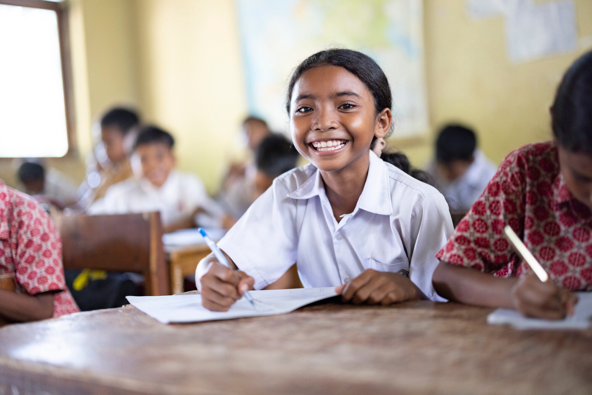 A cheerful young girl in a white school uniform writes in her notebook, surrounded by classmates in a lively classroom.