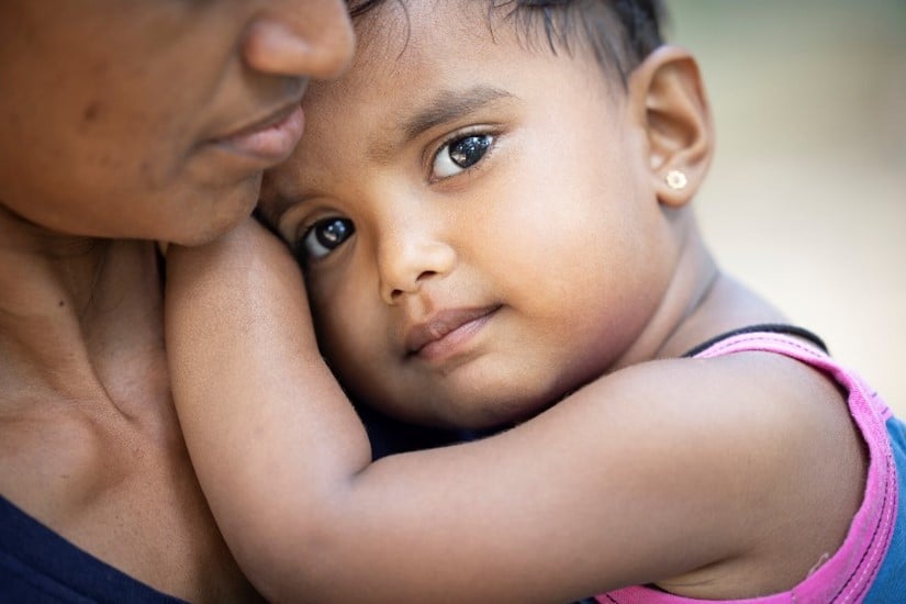A young child with earrings and a pink tank top embraces their mother, gazing softly into the camera while resting on her shoulder.