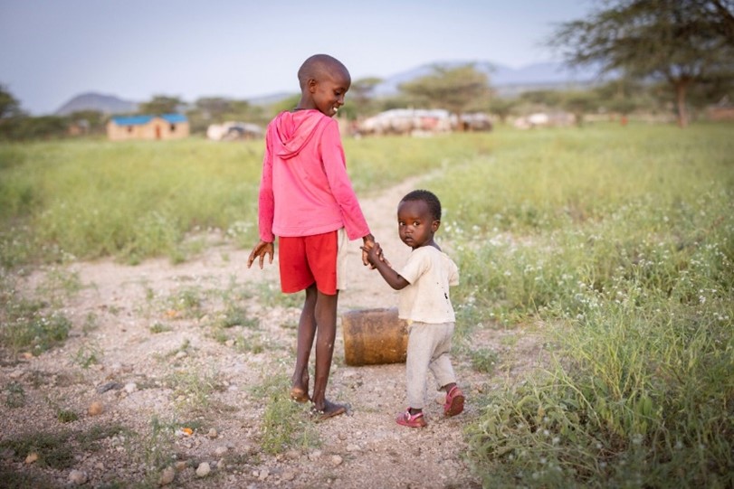 A young boy in a pink hoodie holds the hand of a smaller child while walking along a rural dirt path, with grass, trees, and a village in the background.