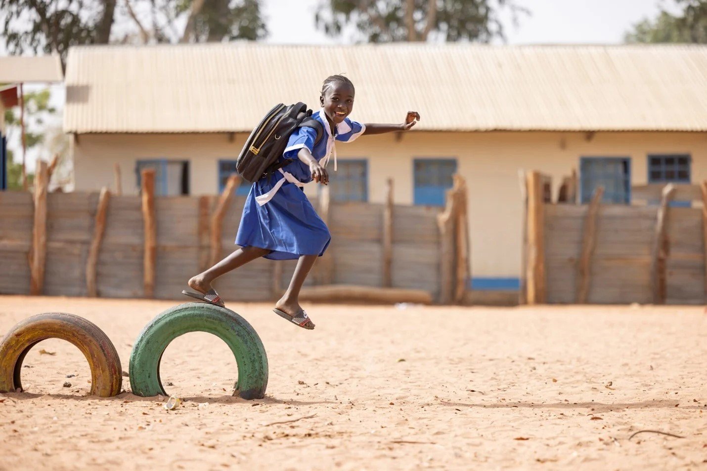A happy schoolgirl in a blue uniform with a backpack balances on tires in a sandy playground in front of a rural school building.