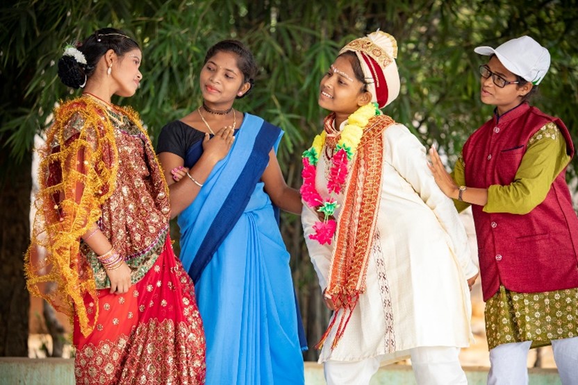 Students in traditional clothing performing a play outdoors, dressed as a bride, groom, and supporting characters, with greenery in the background.