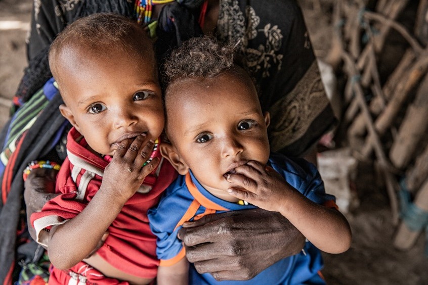 Two babies, one in red and one in blue, sit on their mother’s lap, sucking on their fingers, with the mother’s hands gently holding them.