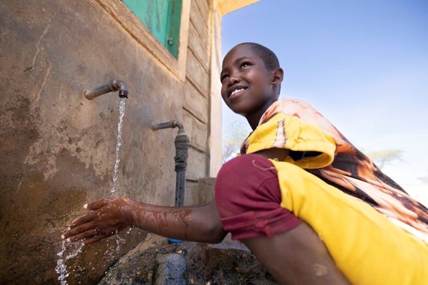 A smiling boy in a yellow and red outfit washes his hands under a running water tap outside a building, with a clear blue sky in the background.