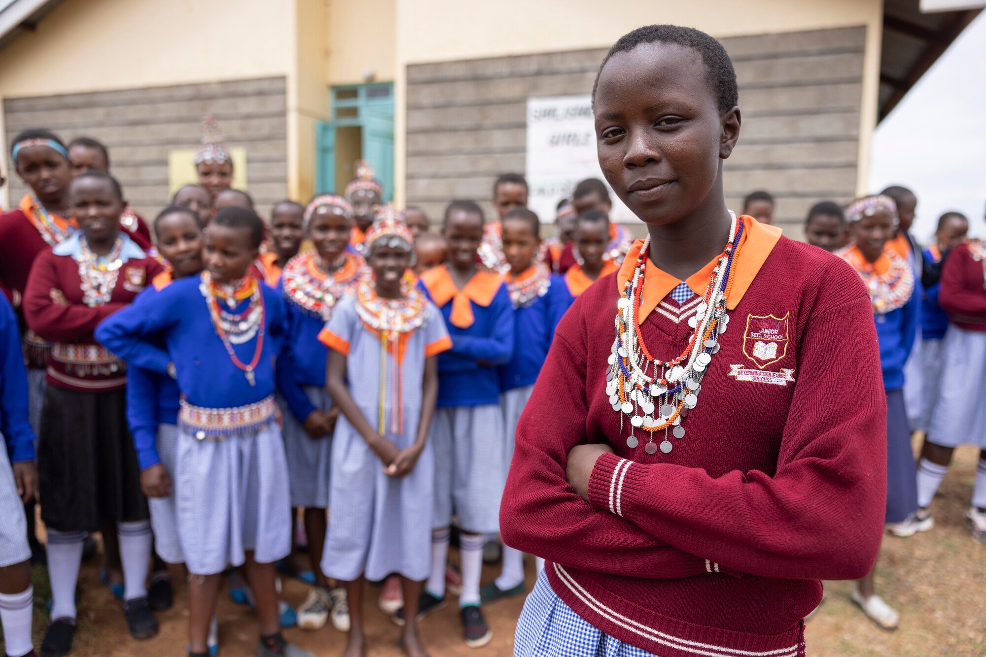 A confident young girl wearing a maroon school uniform and traditional beaded necklaces stands with her arms crossed in front of a group of students outside a school.