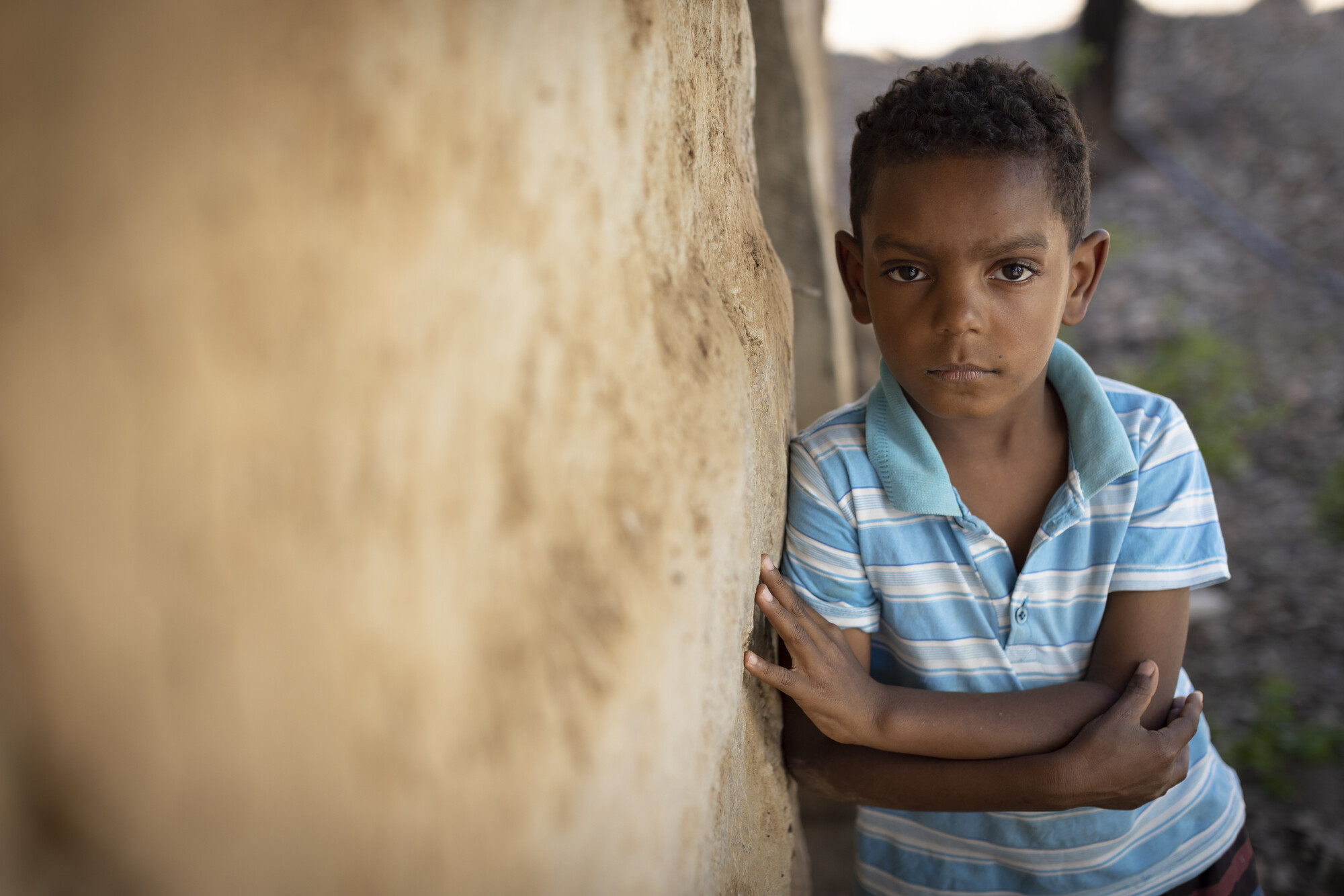 Boy in a blue-striped shirt leans against a mud wall, gazing intently.