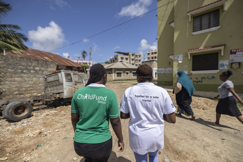 Two women in Kenya wearing ChildFund shirts walk through the streets, their backs facing the camera.