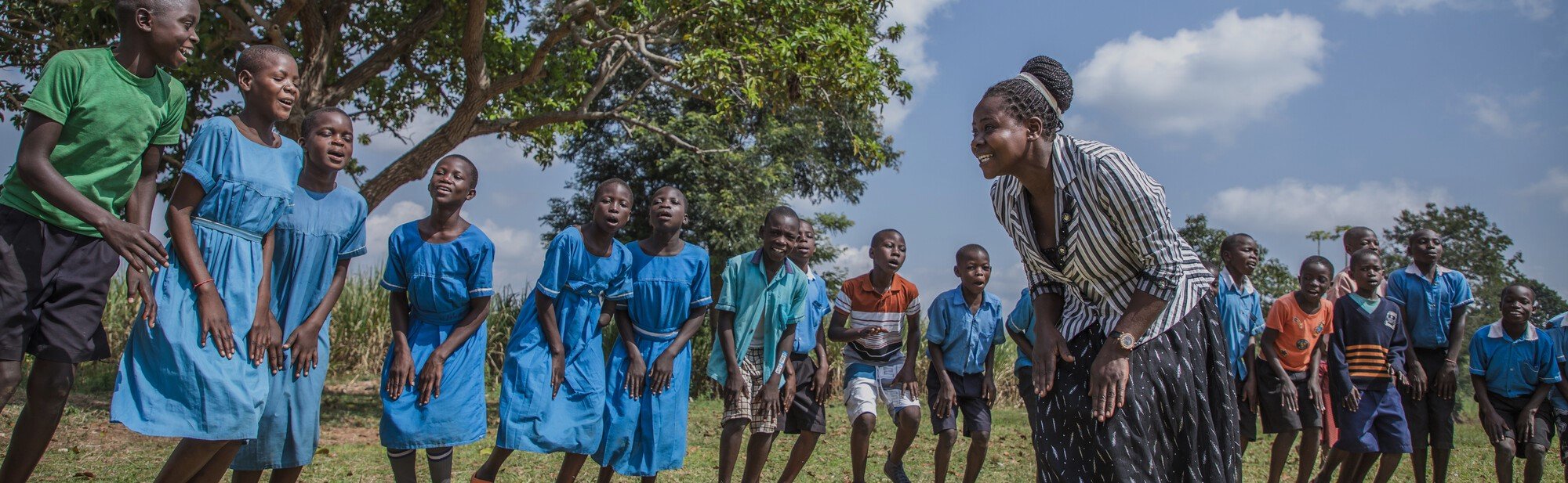Children in blue uniforms laughing and engaging in an outdoor activity with a smiling teacher under a bright sky.