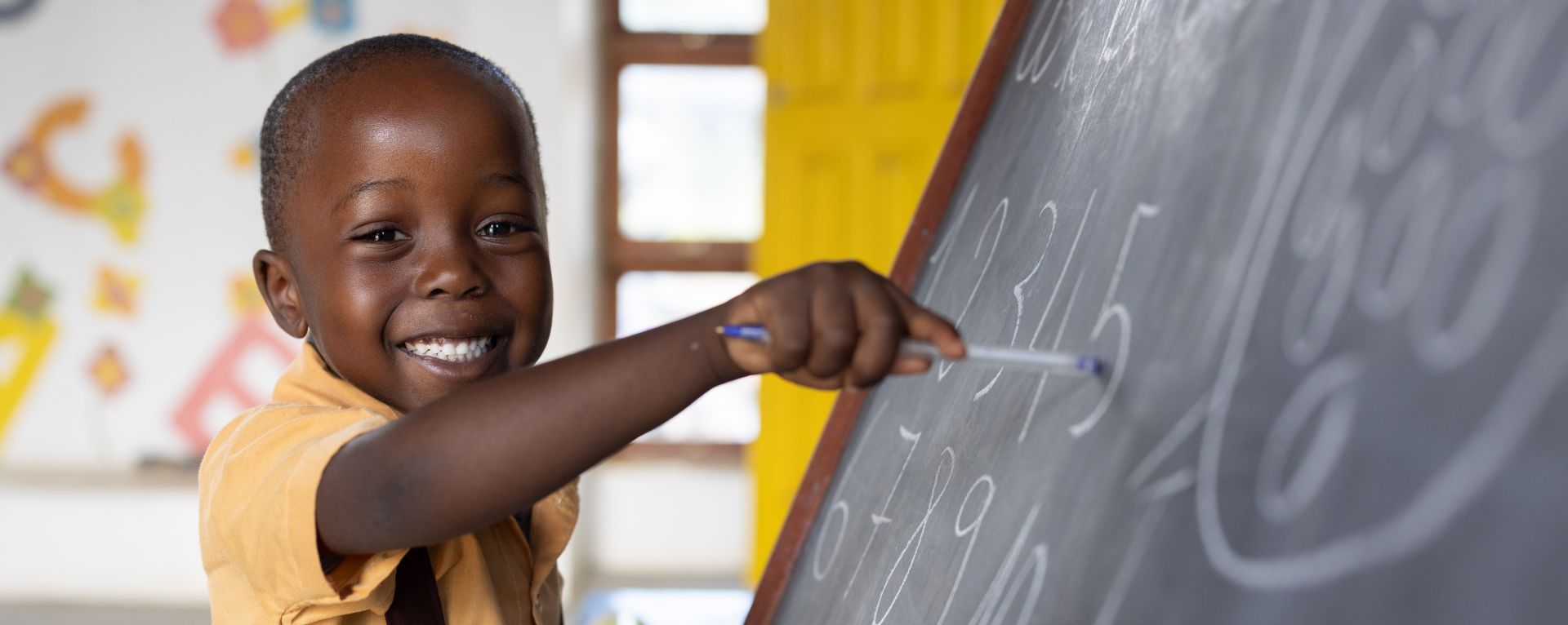 A young boy wearing a yellow shirt smiles while pointing at a chalk board.
