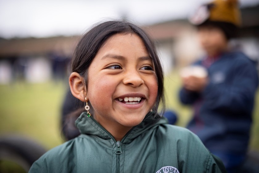 A young girl with dark hair and earrings smiles joyfully while wearing a green jacket, with other children in the background.