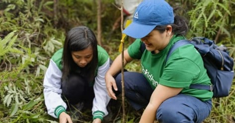 Two women in green shirts plant a sapling in a forested area, smiling as they work.