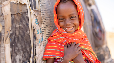 miling young girl in an orange headscarf standing beside a rustic shelter, symbolizing hope and resilience.