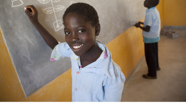 Smiling young girl writing on a chalkboard in a classroom, representing education and opportunity.