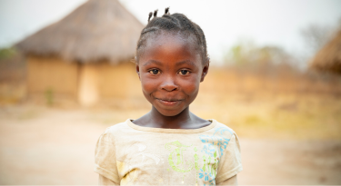 Smiling young girl standing in front of traditional huts, representing resilience and community spirit.