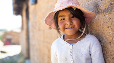 Smiling young girl in a pink sun hat leaning against an adobe wall, representing joy and cultural heritage.