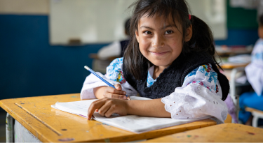 Smiling young girl writing in a notebook at her school desk, representing education and opportunity.