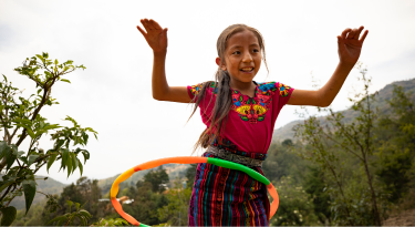 Smiling girl in traditional Guatemalan clothing playing with a hula hoop, representing joy and culture.