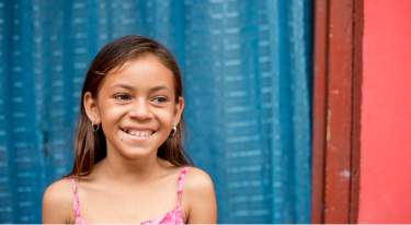 Smiling young girl in a pink dress standing in front of a blue curtain, symbolizing hope and resilience.