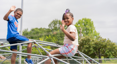 Two children waving while climbing on a jungle gym, symbolizing play and friendship.