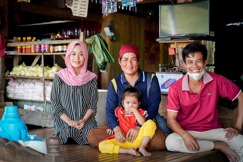 A family in Cambodia sits in front of a small shop, smiling at the camera.