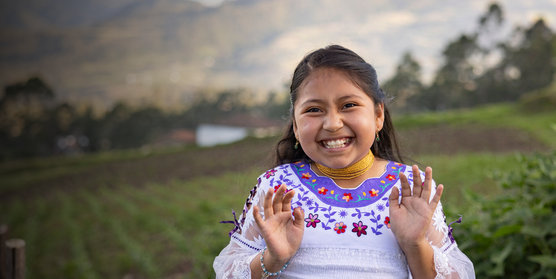 A cheerful young girl in a white and purple embroidered dress raises her hands playfully while standing in a green rural landscape with mountains in the background.